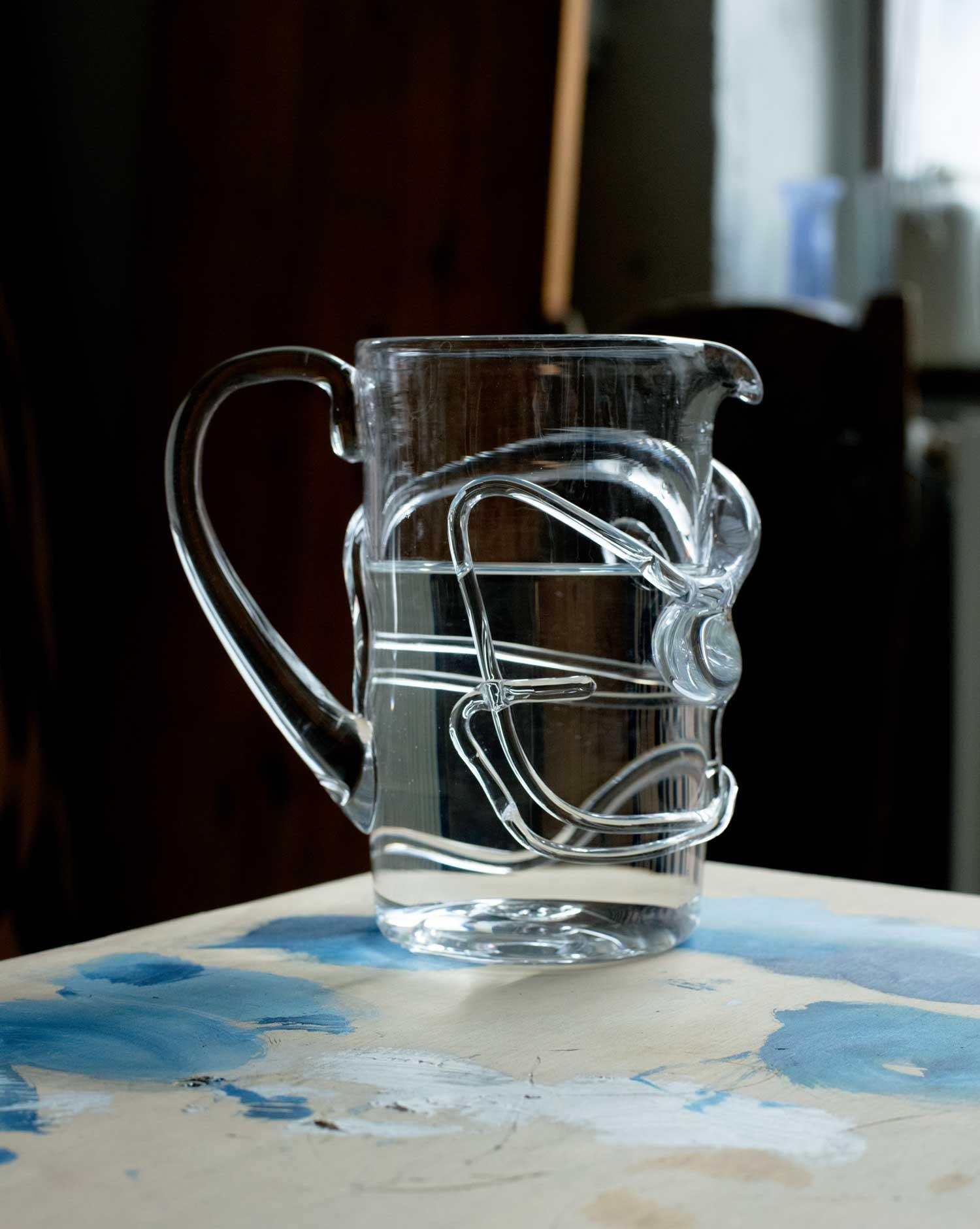 Glass pitcher with melted glass details with handle on the left side with water on a table